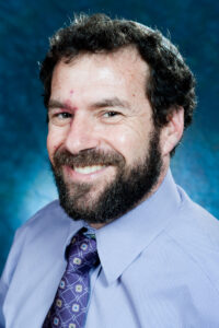A bearded man in a light purple shirt and patterned tie smiles at the camera, set against a blue background.