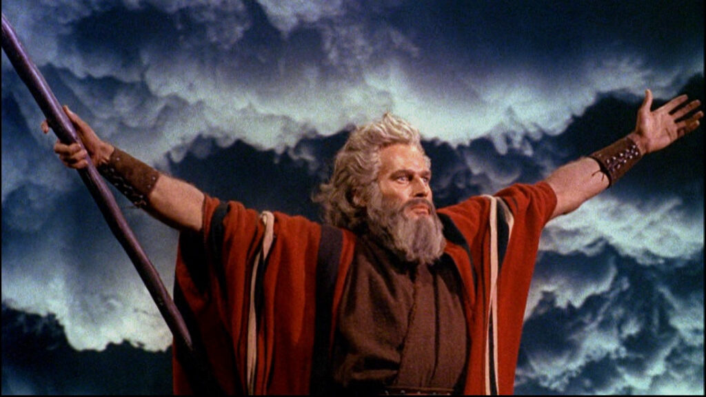 Charlton Heston as Moses, arms spread wide, holding his staff as the sky storms.