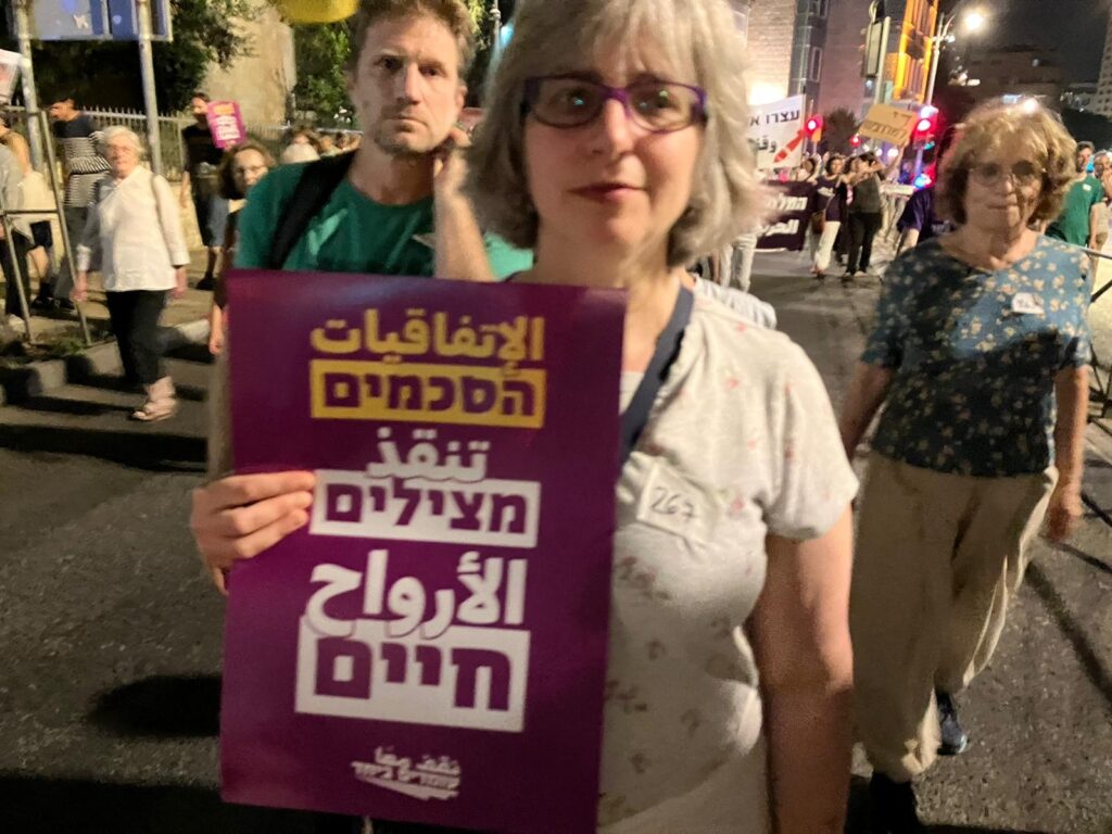 Rabbi Mira Wasserman, Ph.D., holding a sign that says in Hebrew, "Agreement saves lives" from the organization Standing Together. Other signs at the Jerusalem march on Saturday night were for the return of the hostages, and end to the war and new elections.