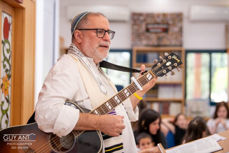 Rabbi Steve Burnstein, wearing tallit and a kippa, strums his guitar. He appears to be in a classroom as children are looking on.