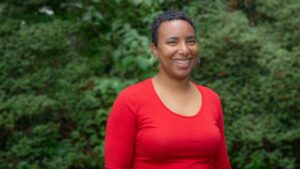 Smiling person in a red shirt standing in front of green foliage.