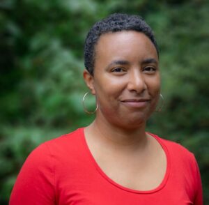 Person with short hair in a red shirt smiles slightly, standing in front of a blurred green background.