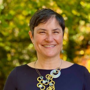 Smiling person with short hair and a statement necklace stands outdoors against a backdrop of greenery.