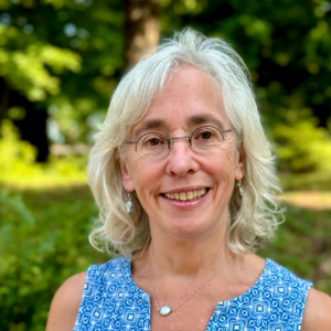 A woman with glasses and gray hair smiles outdoors, wearing a blue patterned top.