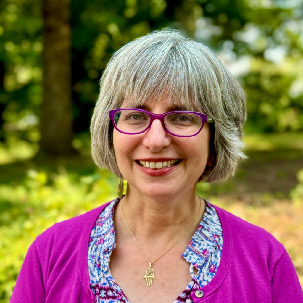 Rabbi Mira Wasserman, with gray hair and purple glasses, smiles outdoors, wearing a purple top and a necklace.