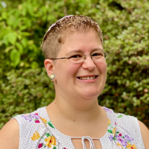 A smiling person with short hair and glasses, wearing a floral top, stands in front of green foliage.