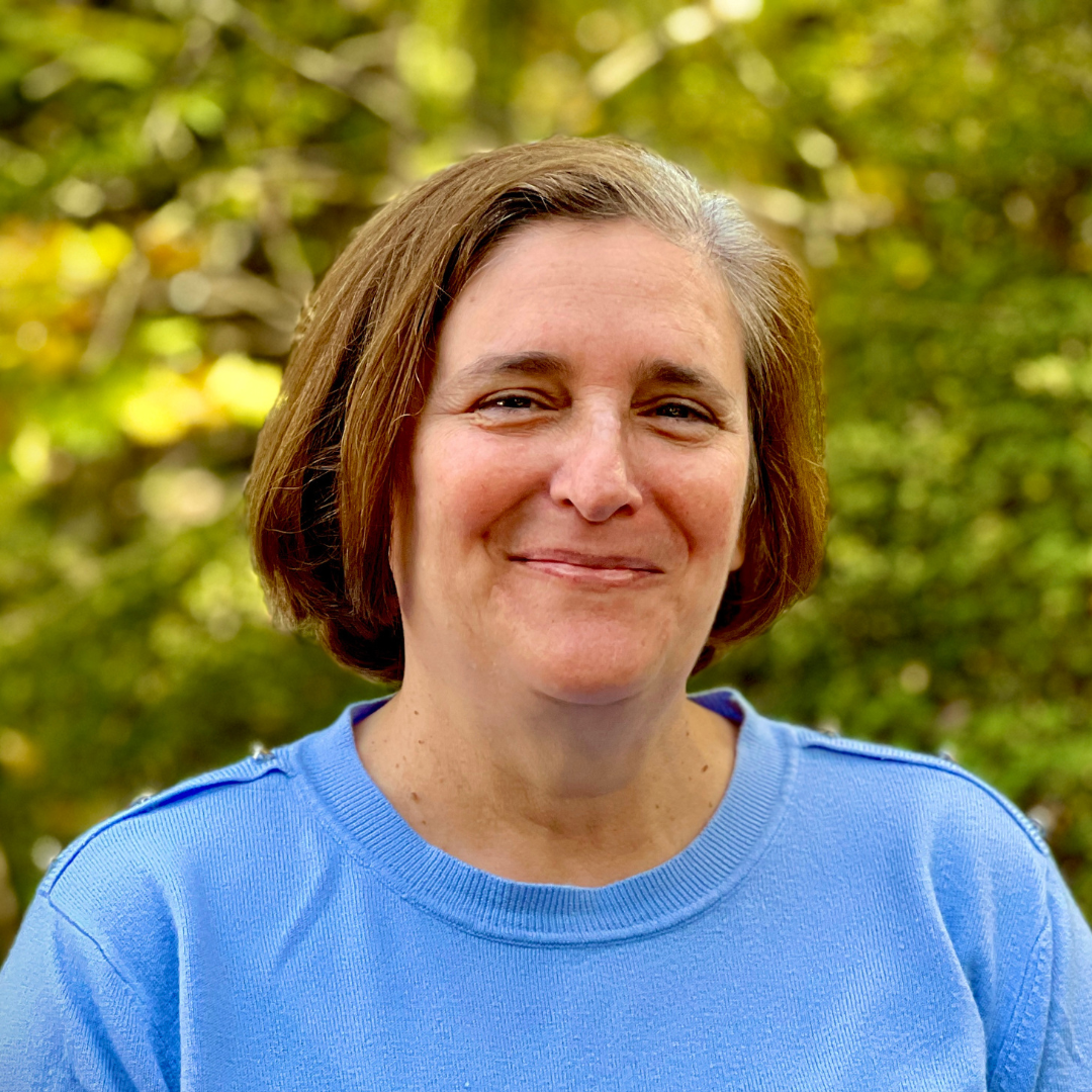 Smiling person with short hair wearing a blue sweater, standing outdoors with green foliage in the background.
