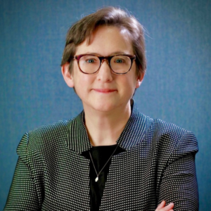 Rabbi Sharon Kleinbaum, sporting short hair and glasses, is wearing a patterned blazer, smiling against a blue background.