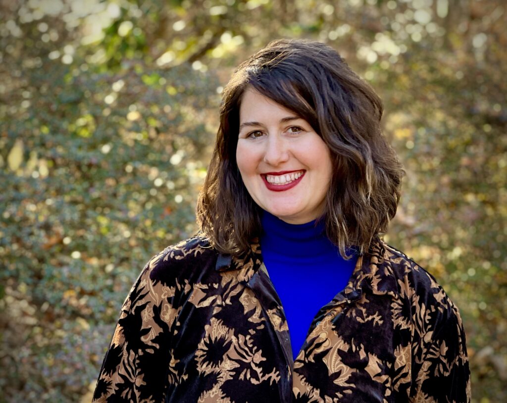 Alana Krivo-Kaufman, with dark hair, wearing a floral jacket and blue top, smiles outdoors with trees in the background.