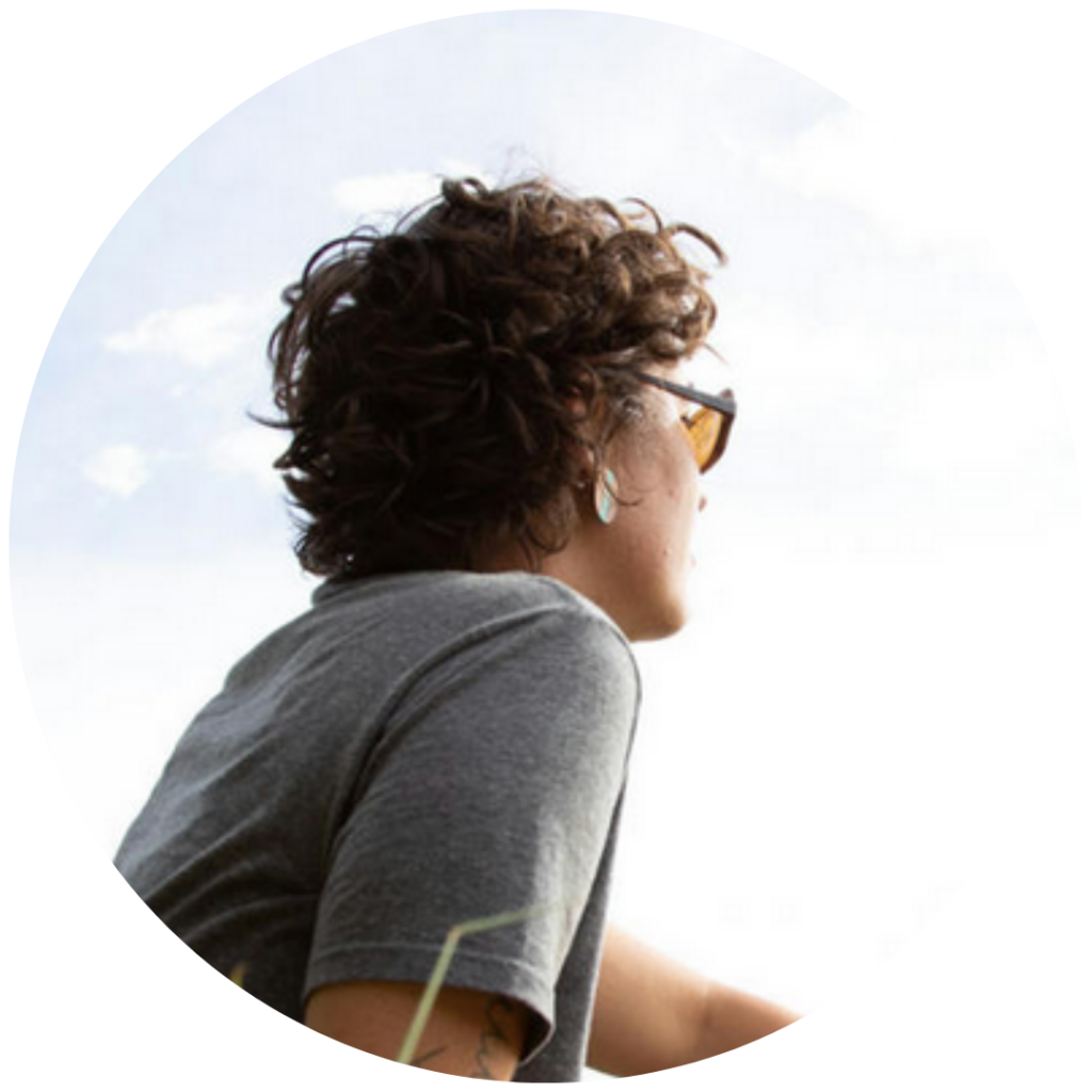 Person with curly hair and sunglasses looking at the sky, wearing a gray t-shirt.