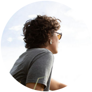 Person with curly hair and sunglasses looking at the sky, wearing a gray t-shirt.