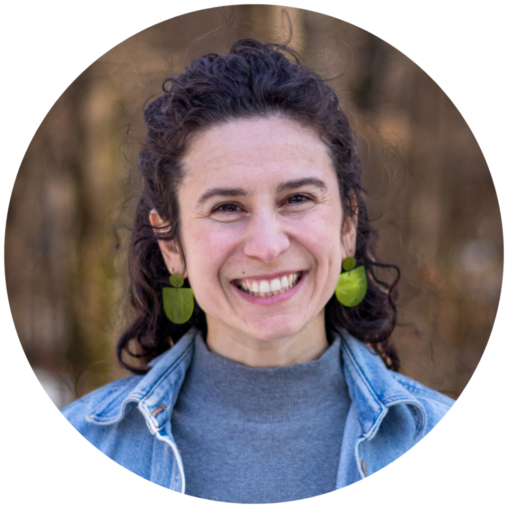 Smiling person with curly hair, wearing a denim jacket and green earrings, standing outdoors.