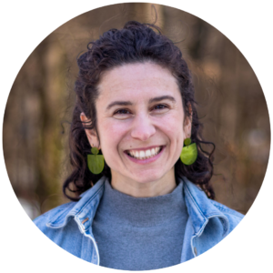 Smiling person with curly hair, wearing a denim jacket and green earrings, standing outdoors.