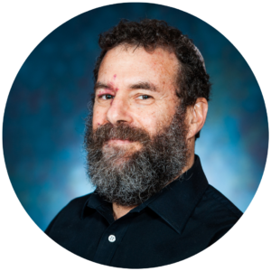 Person with a beard wearing a dark shirt, standing against a blurred blue background.