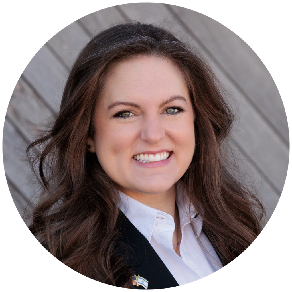 Smiling woman with long brown hair, wearing a suit and white shirt, against a wooden background.