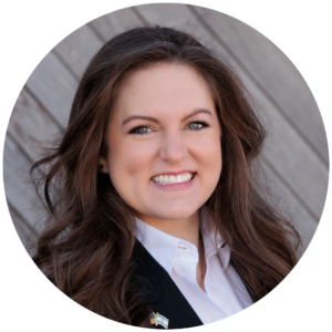 Smiling woman with long brown hair, wearing a suit and white shirt, against a wooden background.