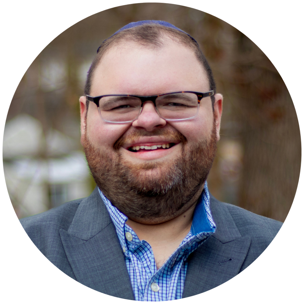 Smiling man with glasses and beard, wearing a suit and checkered shirt, in a blurred outdoor background.