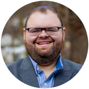 Smiling man with glasses and beard, wearing a suit and checkered shirt, in a blurred outdoor background.