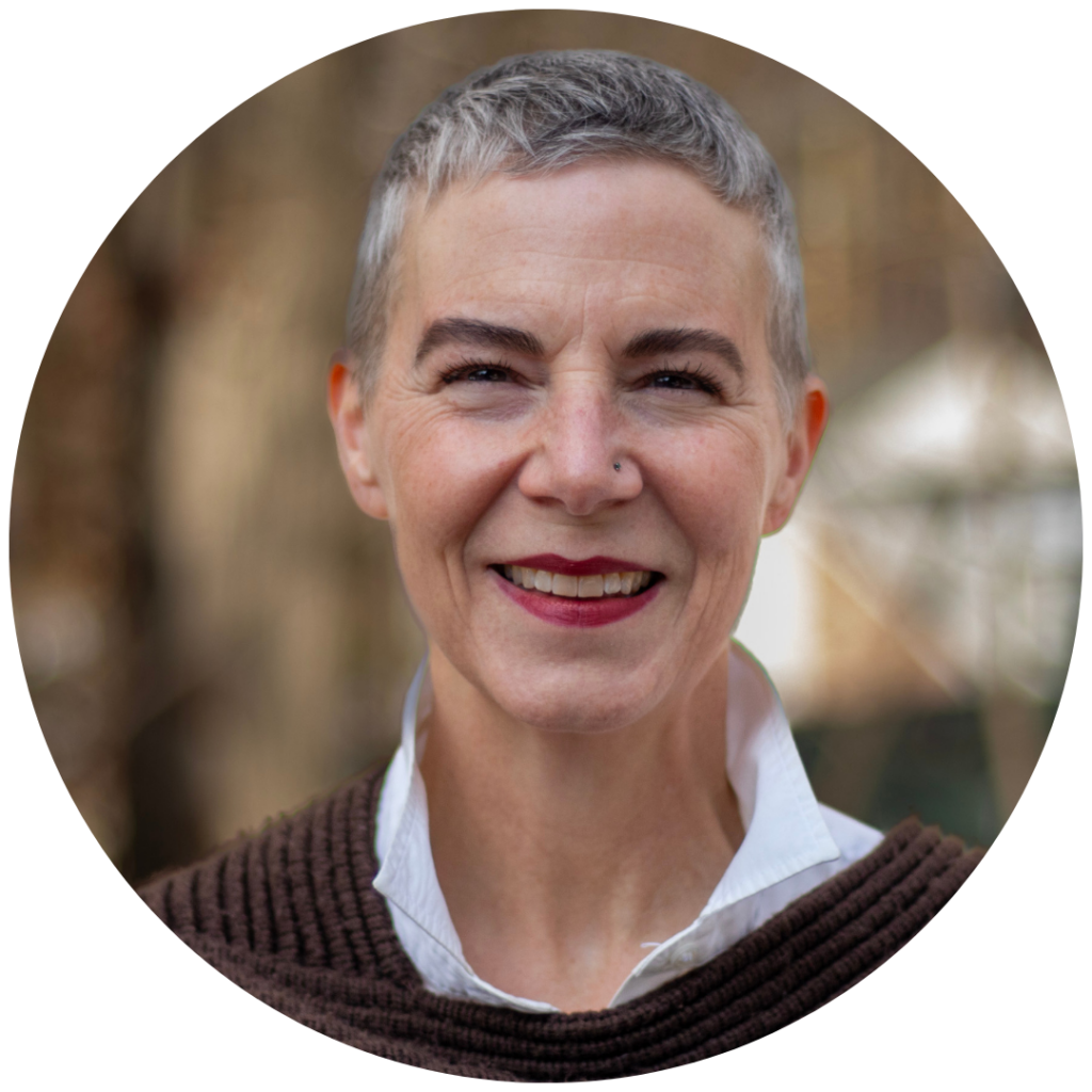 Smiling woman with short gray hair and a brown sweater, standing outdoors with a blurred background.