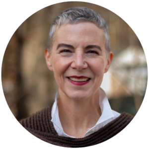 Smiling woman with short gray hair and a brown sweater, standing outdoors with a blurred background.