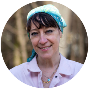 Woman with a blue headscarf smiling outdoors, wearing a pink shirt and layered necklaces.