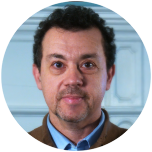 Man with curly hair and goatee wearing a blue shirt and brown sweater, standing indoors against a light background.