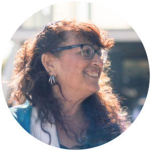 Smiling woman with glasses and curly hair, wearing a white blouse and blue jacket, in outdoor setting.