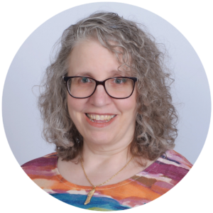 A smiling woman with curly gray hair and glasses, wearing a colorful top and necklace, on a plain background.