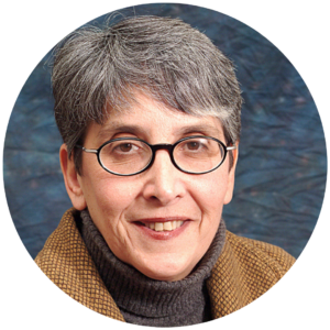 Person with short gray hair, glasses, and a brown jacket smiles against a blue background.