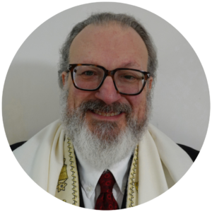 A smiling person with glasses, a beard, and wearing a white garment with gold symbols on the edges.
