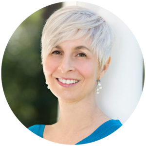 Smiling person with short white hair and earrings, wearing a blue top, standing against a light background.