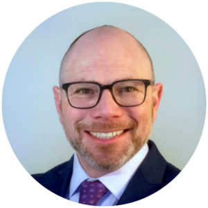 Man with glasses and a beard smiling in a suit and tie against a plain background.