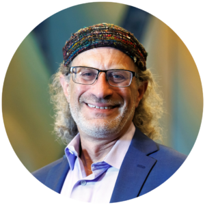 Smiling man with glasses and a colorful woven cap, wearing a suit jacket and shirt.