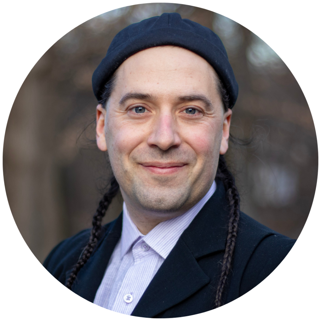 Smiling person with long braided hair and a black hat, wearing a black jacket and light shirt, outdoors.