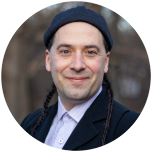 Smiling person with long braided hair and a black hat, wearing a black jacket and light shirt, outdoors.