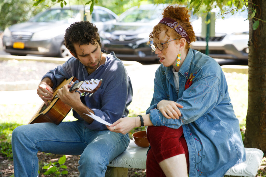 Two people sitting outside; one plays guitar while the other holds sheet music. They seem focused and relaxed.