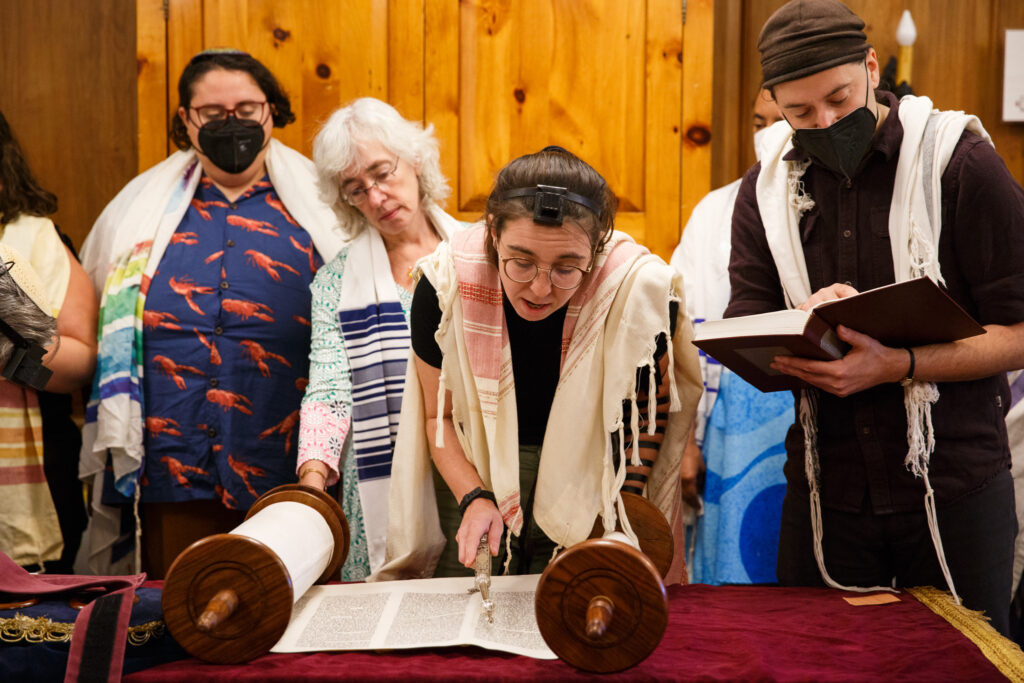 People in prayer shawls read from the Torah, one holding a yad pointer, in a wood-paneled room.
