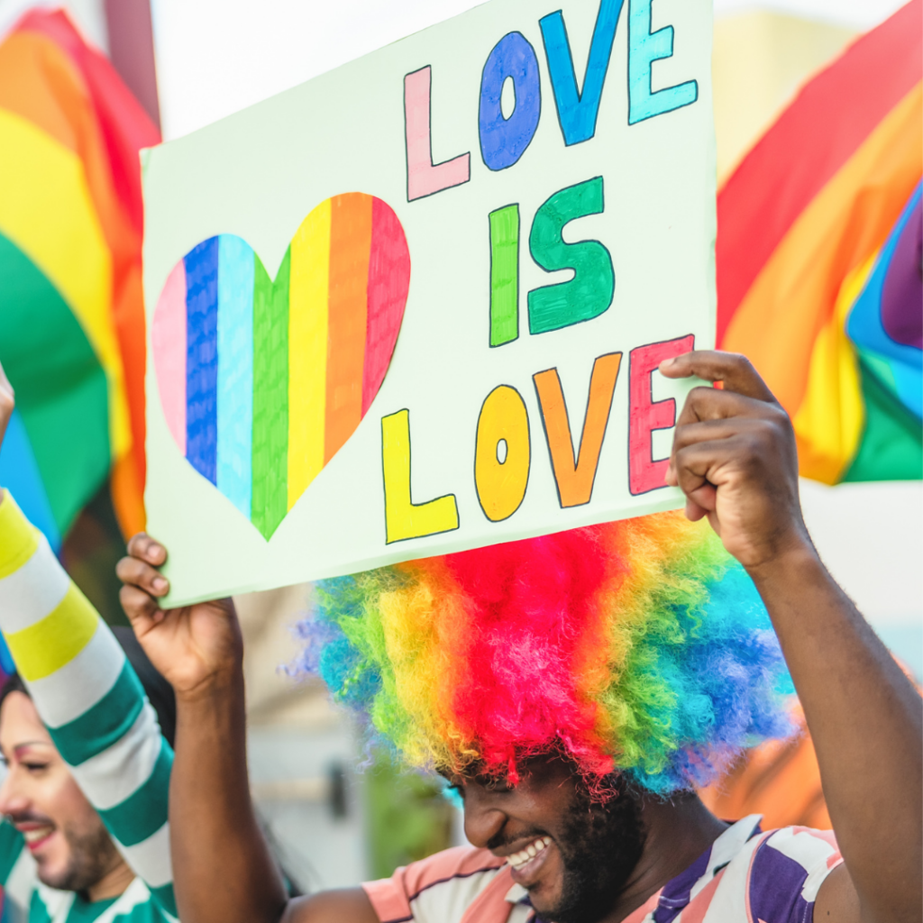 Person in a rainbow wig holding a “Love is Love” sign with a rainbow heart at a pride event.
