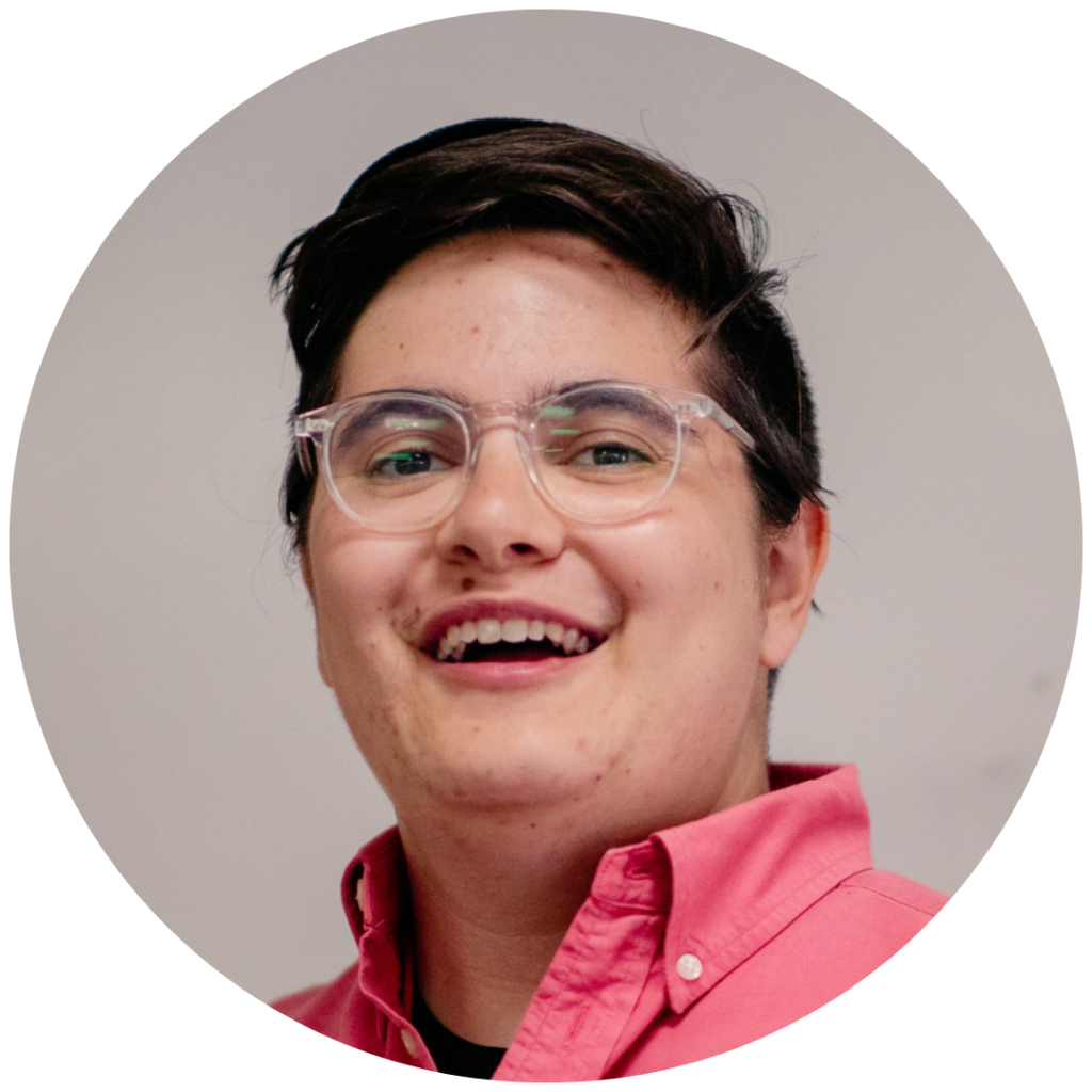 Person with short dark hair, clear glasses, and a pink shirt smiling at the camera against a plain background.