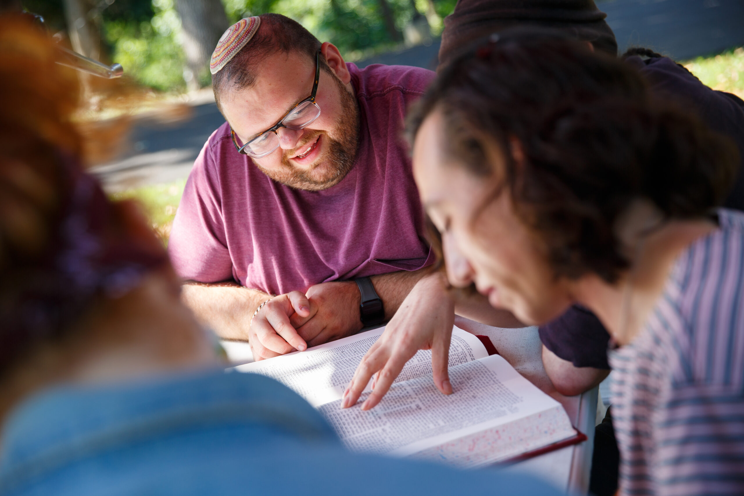 A group of people studies a book outdoors, one person points at the text while others look on.