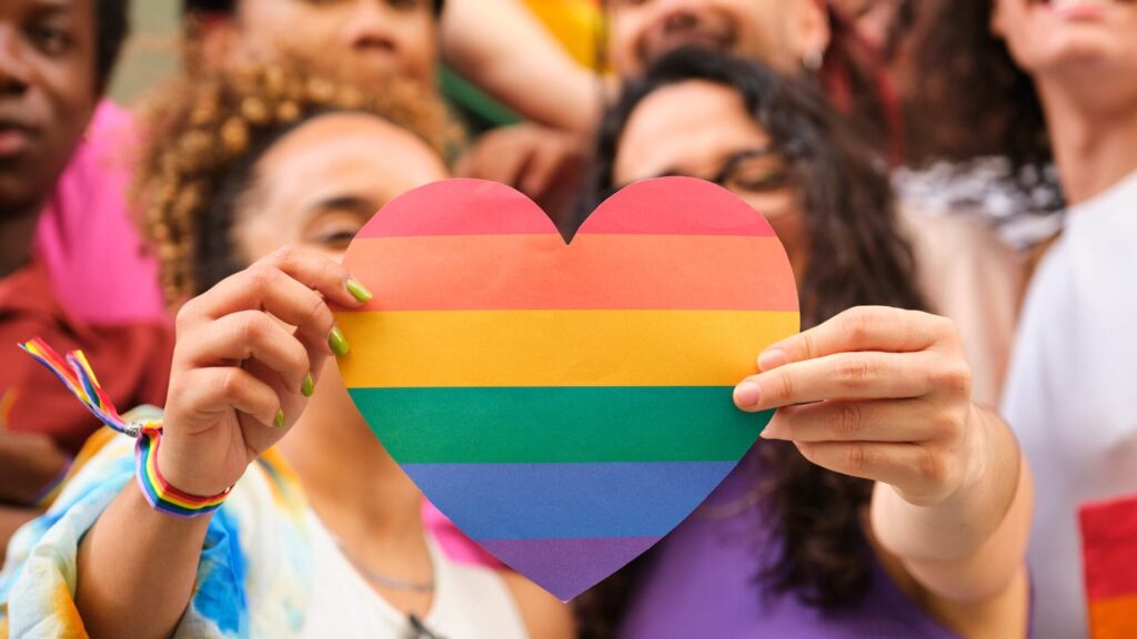Two people hold a rainbow heart sign, surrounded by a diverse, smiling group at a pride event.