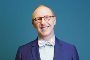 Smiling man in glasses and a bow tie wearing a blue suit, posing in front of a solid blue background.