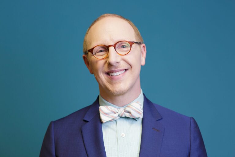 Smiling man in glasses and a bow tie wearing a blue suit, posing in front of a solid blue background.