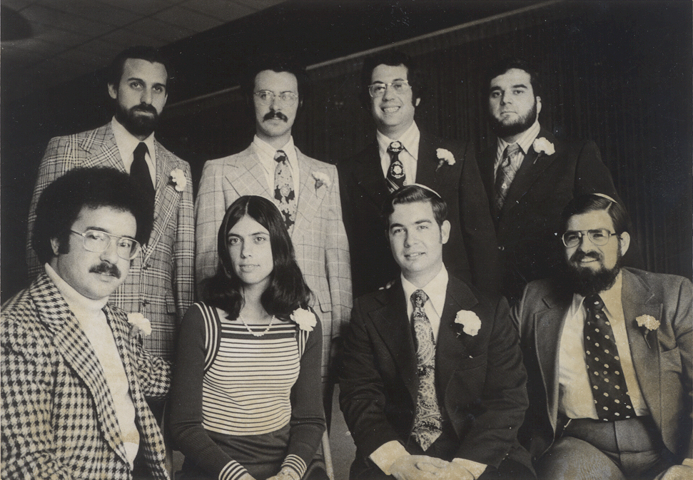 Eight people in formal 1970s attire, posing for a group photo indoors, some wearing boutonnieres.