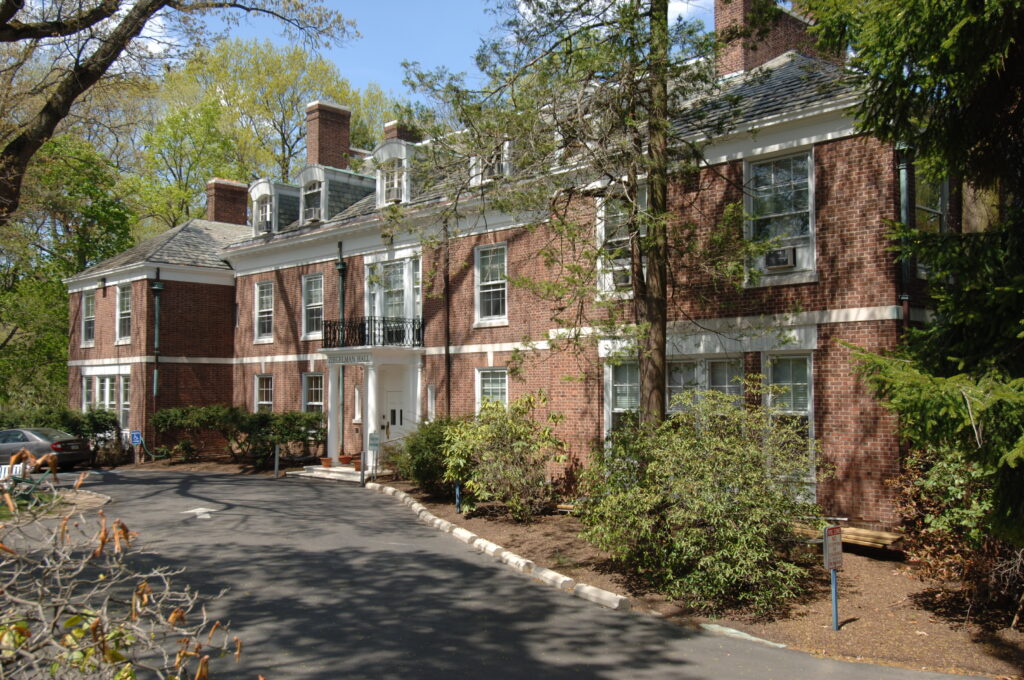 Large brick building with white trim, surrounded by trees and shrubs, along a curved driveway.