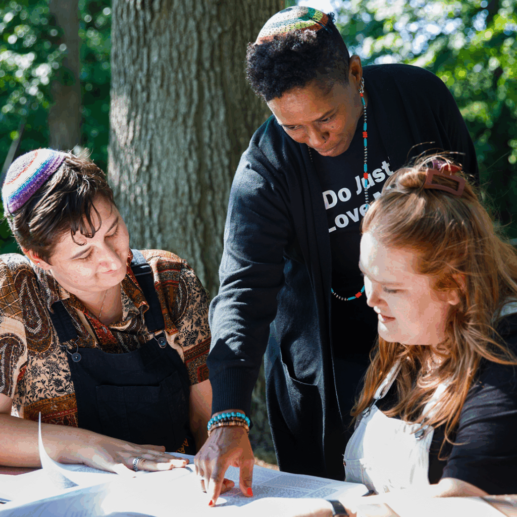 Three people wearing yarmulkes study together outdoors, looking at an open book while one points at the page.