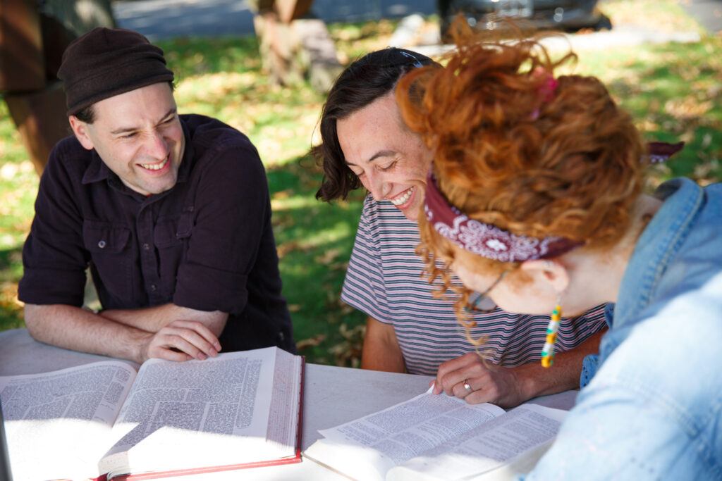 Three people sit at an outdoor table, smiling and studying large open books together.