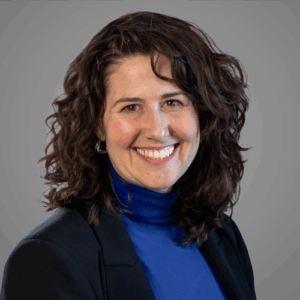 Woman with curly brown hair in a blue turtleneck and dark blazer, smiling against a plain gray background.