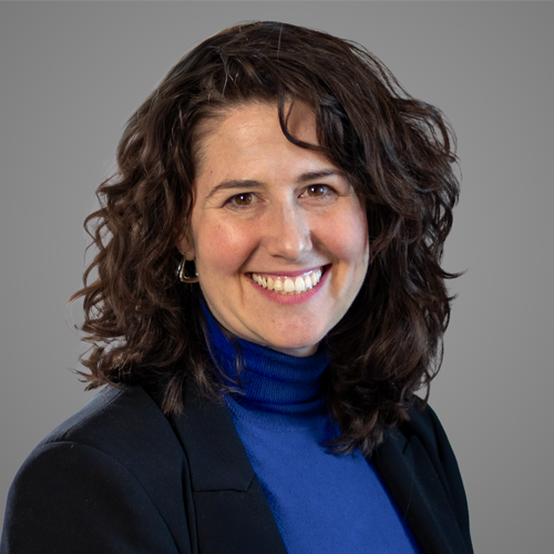 Woman with curly brown hair in a blue turtleneck and dark blazer, smiling against a plain gray background.