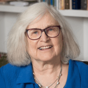 Elderly woman with white hair, glasses, and a blue shirt smiling in front of shelves with books.